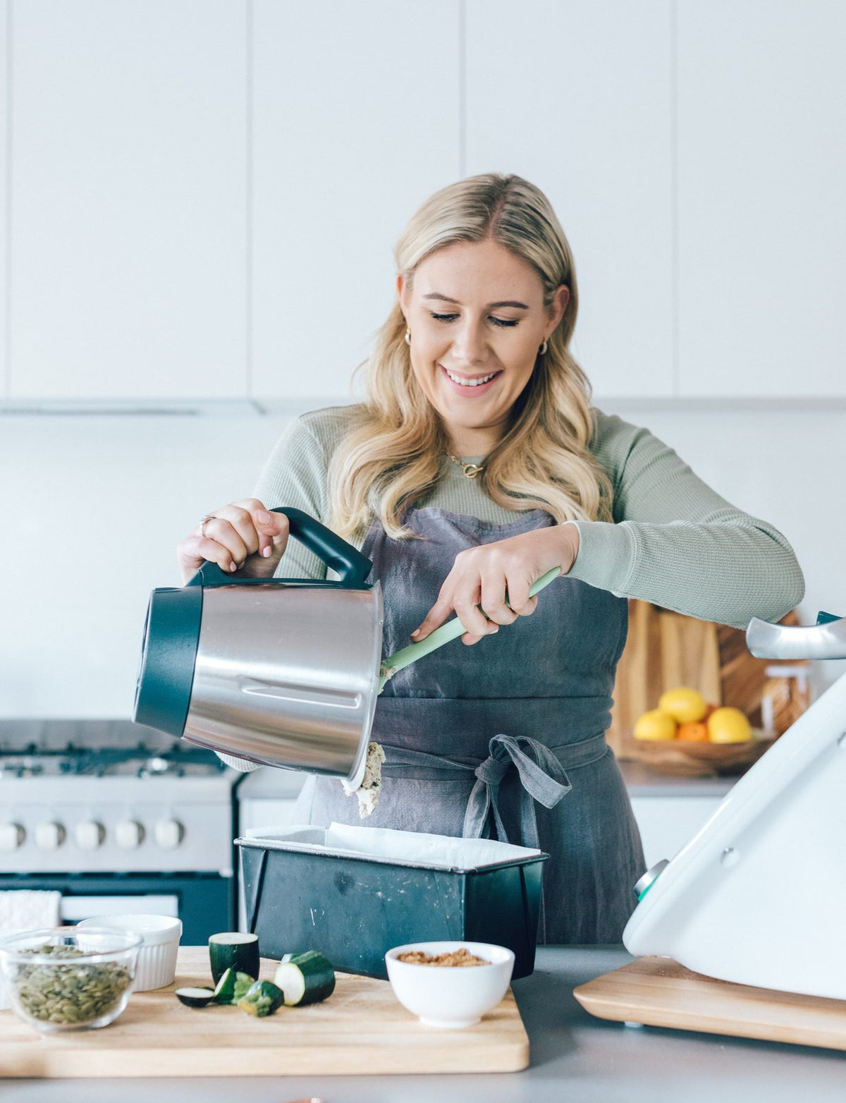 Alyce tipping zucchini loaf batter from a Thermomix bowl into a lined loaf tin, easing it out with a green Thermo Spoon Spatula; the accessories for Thermomix machines she uses every day