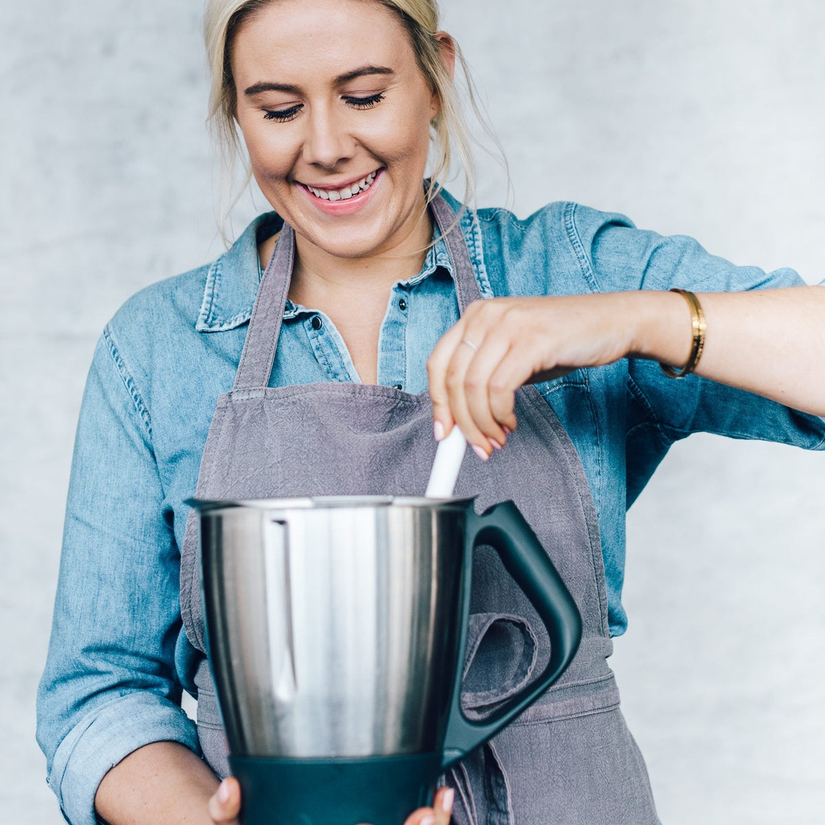 Alyce in a denim shirt and grey apron, smiling as she reaches into a stainless steel Thermomix mixing bowl with a white silicone Thermo Spoon Spatula; what is safe to use inside the bowl