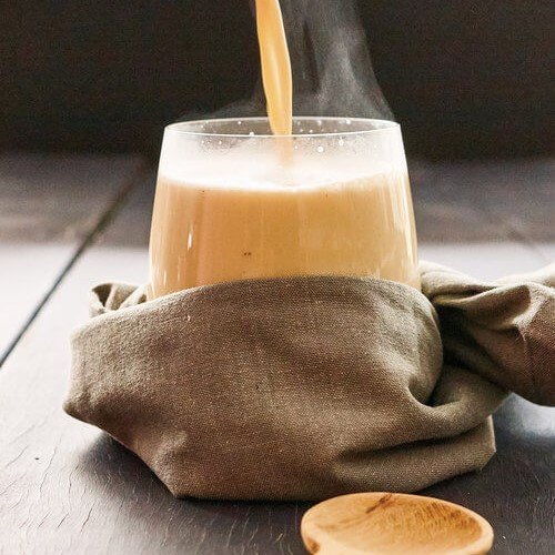 Spiced caramel latte made in the Thermomix being poured steaming into a glass tumbler wrapped in a linen napkin, on a dark timber table with a small wooden spoon alongside