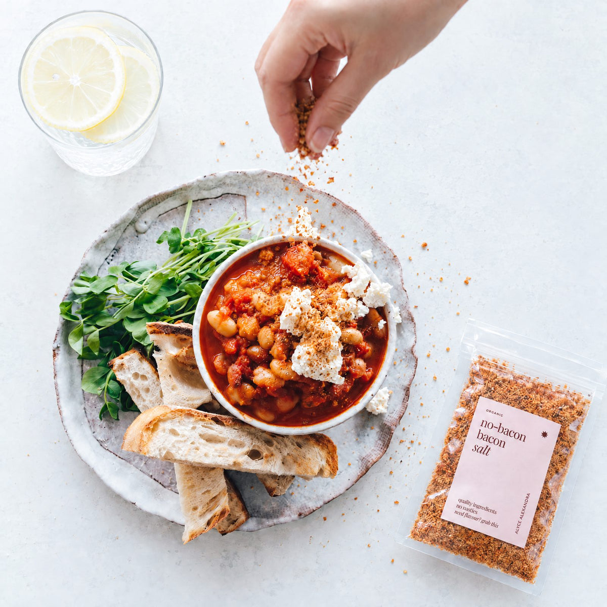 Hand sprinkling No-Bacon Bacon Salt over a bowl of tomato baked beans topped with crumbled white cheese, served with toasted bread and pea shoots beside a packet of the salt; bacon flavour without bacon, no frying needed