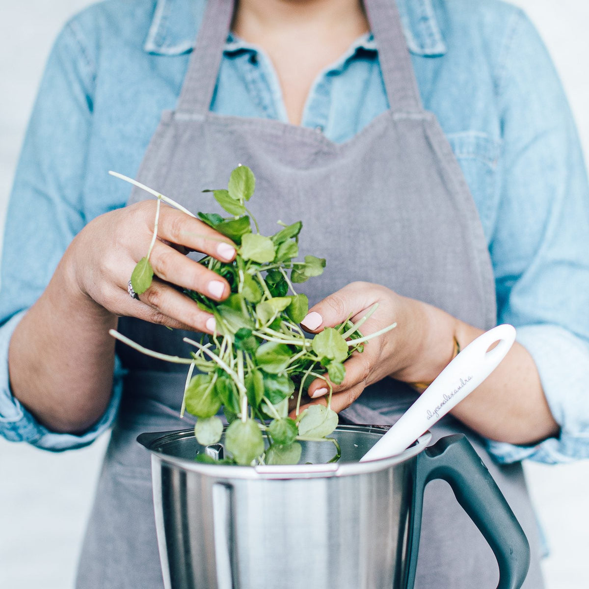 Hands dropping a handful of fresh watercress into a Thermomix bowl, with a Thermo Spoon Spatula resting on the lid; how to stop a green smoothie coming out gritty