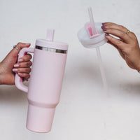 One hand holding the blush pink 1L Insulated Tumbler while the other swaps in the second interchangeable lid with its straw