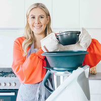 Woman in an apron and oven mitts lifting a stack of steamer basins beside a Thermomix on the benchtop