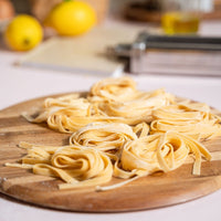 Nests of fresh fettuccine resting on a wooden board with lemons and the pasta roller in the background