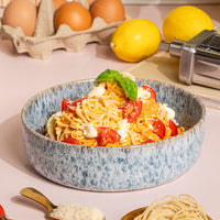 Bowl of spaghetti with cherry tomatoes, feta and basil on a pink bench, with eggs, lemons and the pasta roller alongside