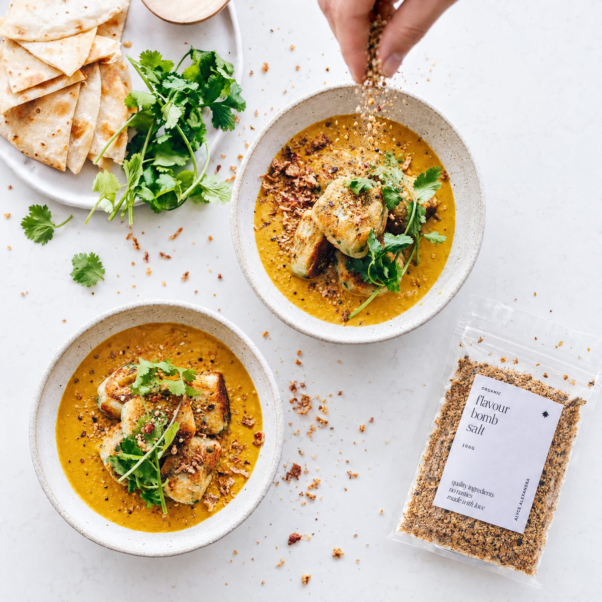 Hand sprinkling Organic Flavour Bomb Salt over two bowls of golden curry with pan-fried dumplings and fresh coriander, beside flatbread and a 100g packet of the salt