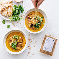Hand sprinkling Organic Flavour Bomb Salt over two bowls of golden curry with pan-fried dumplings and fresh coriander, beside flatbread and a 100g packet of the salt