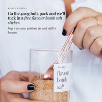 Hands holding a glass jar of Organic Flavour Bomb Salt with a copper spoon; go the 400g bulk pack and a free Flavour Bomb Salt sticker is tucked in, so you can pop it on your prettiest jar and refill it forever