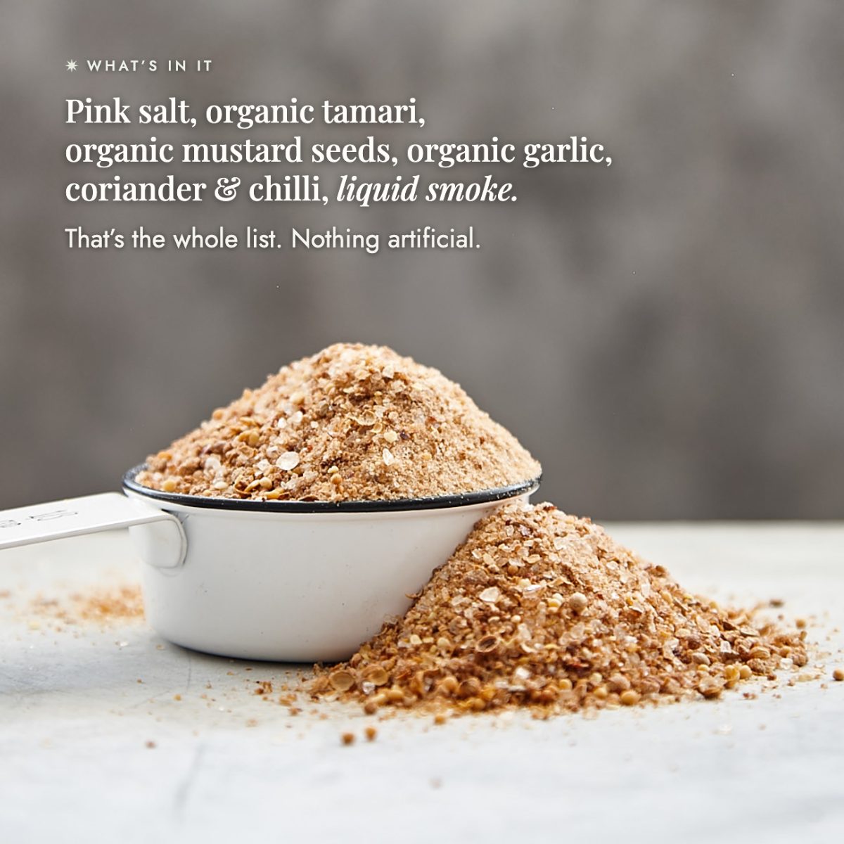 Mound of Organic Flavour Bomb Salt spilling from a measuring cup showing the whole ingredient list: pink salt, organic tamari, organic mustard seeds, organic garlic, coriander and chilli, and liquid smoke; that is the whole list, nothing artificial