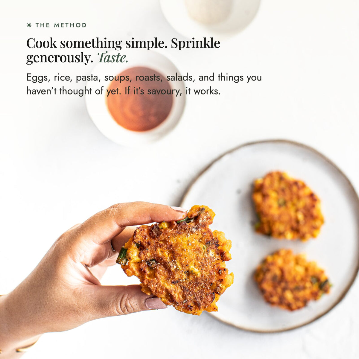 Hand holding a golden corn fritter dusted with Organic Flavour Bomb Salt beside a plate of fritters and dipping sauce; cook something simple, sprinkle generously and taste, on eggs, rice, pasta, soups, roasts and salads