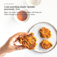 Hand holding a golden corn fritter dusted with Organic Flavour Bomb Salt beside a plate of fritters and dipping sauce; cook something simple, sprinkle generously and taste, on eggs, rice, pasta, soups, roasts and salads