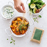 Hand sprinkling Organic No-Chicken Chicken Salt over a bowl of potato curry topped with microgreens, beside cucumber raita, fresh mint, lime and a 100g packet of the salt