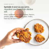 Hand holding a golden corn fritter beside a plate of fritters; sprinkle No-Chicken Chicken Salt over as you serve, anywhere you would reach for chicken salt, on chips, cauliflower, corn and chickpeas, popcorn, pretzels, potato wedges and pumpkin, fritters, fried rice, fish and frittata