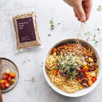 Hand sprinkling Bolognese Boost Salt over a big bowl of spaghetti bolognese with cherry tomatoes and microgreens, beside a packet of the salt