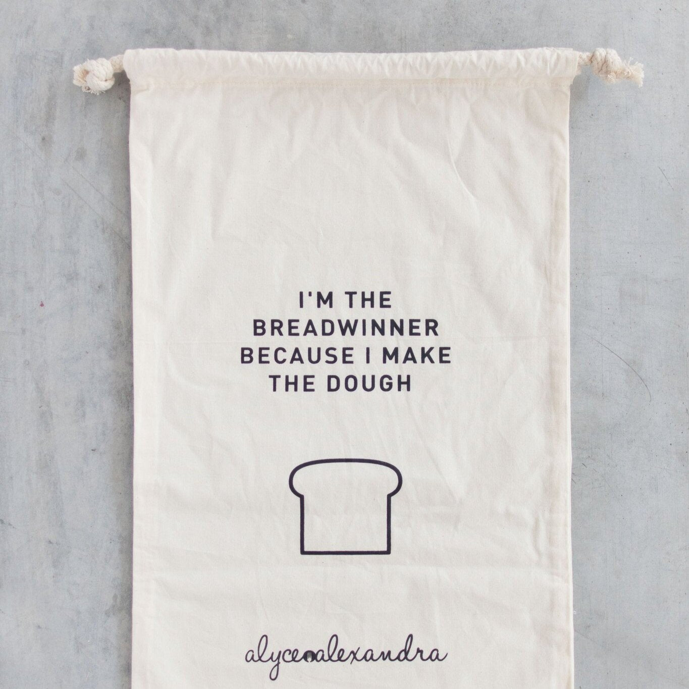Breadwinner Bread Bag in natural organic cotton with a drawstring top, printed with a loaf illustration and the line I'm the breadwinner because I make the dough