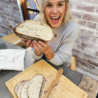 Alyce holding up a large round sourdough loaf of bread cut in half, with the Breadwinner Bread Bag and a bread board on the table