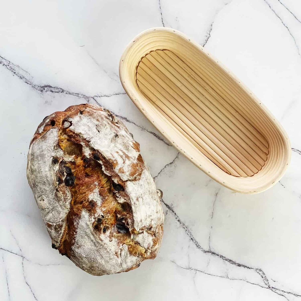 Fruit sourdough loaf of bread beside an oval rattan proving basket on marble