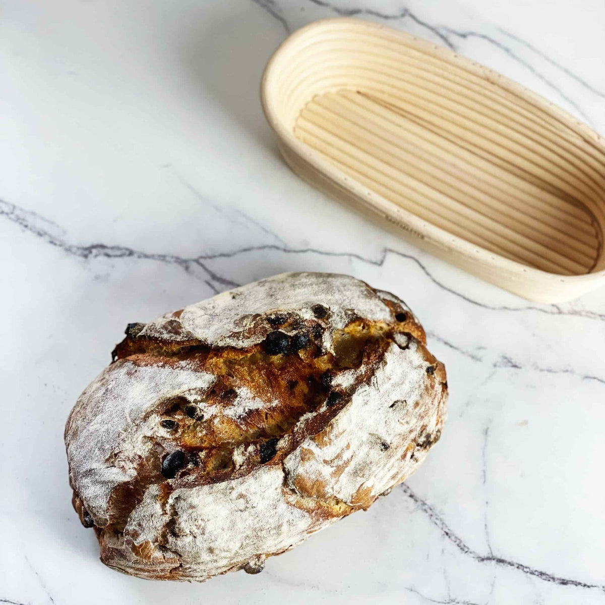 Fruit sourdough loaf of bread and an oval proving basket on a marble bench