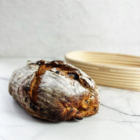 Fruit sourdough loaf of bread beside its oval proving basket