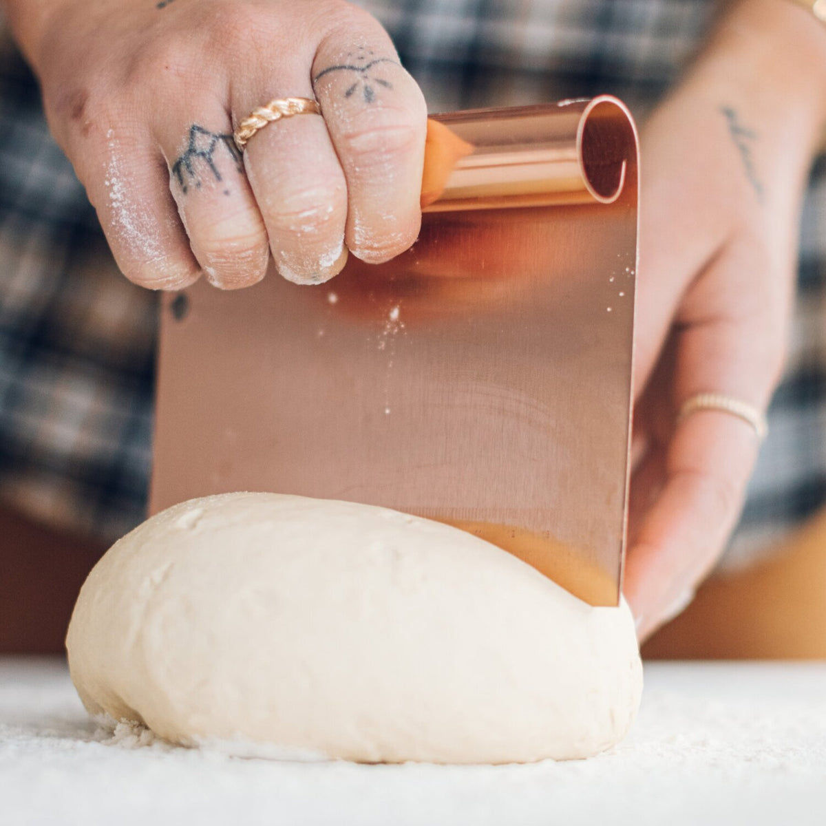 Copper Dough Scraper held blade down against a round of dough, showing the etched measurement markings