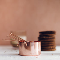Copper Measuring Cups stacked beside a candle and a pile of biscuits on a bench