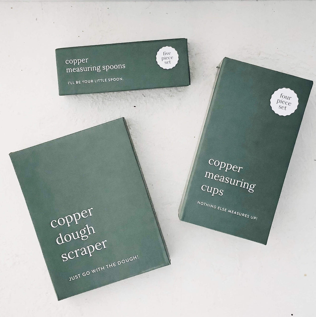 Copper Measuring Cups and Copper Dough Scraper in their dark green gift boxes, laid flat on a pale bench