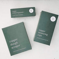 Copper Measuring Cups and Copper Dough Scraper in their dark green gift boxes, laid flat on a pale bench