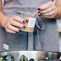 Hand holding a jar of preserved lemons labelled by hand, with a Copper Measuring Spoon on the bench alongside