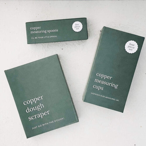Copper Measuring Spoons and Copper Measuring Cups in their dark green gift boxes, laid flat on a pale bench