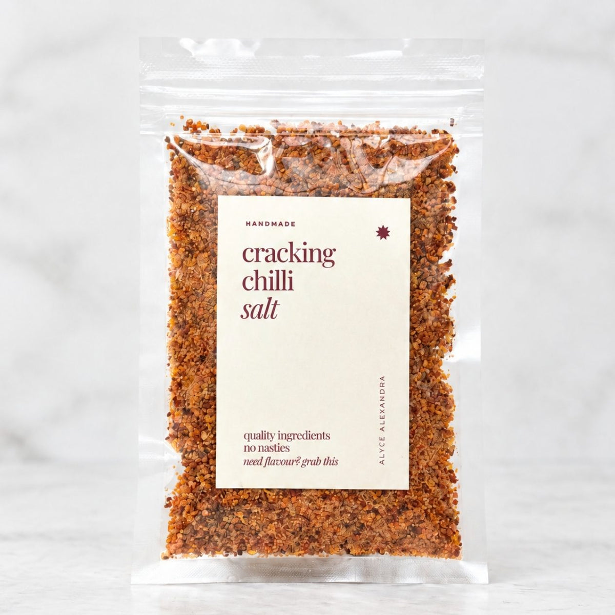 Alyce's Cracking Chilli Salt in a clear 100g packet with a cream label and deep red lettering, the coarse orange-red chilli salt visible through the front