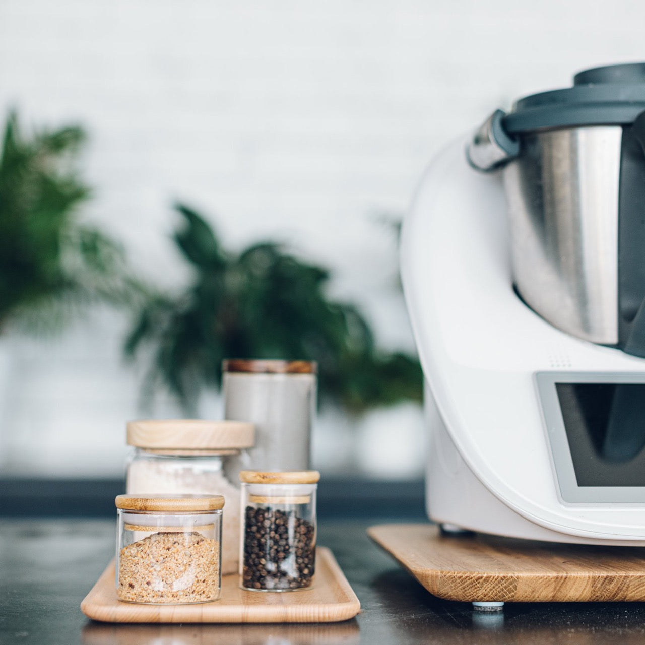 Thermomix sitting on the wooden Deluxe Tray on a benchtop, with glass storage jars and plants alongside