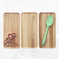 Three wooden Deluxe Trays side by side holding copper measuring spoons and a green spatula, showing the timber grain variations
