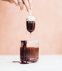 Hand dipping an ice cream into a glass of melted dark chocolate on a pink background