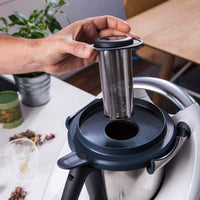 Hand lowering the Filtix Thermo Tea Filter into the lid of a Thermomix, with loose leaf tea and a glass alongside