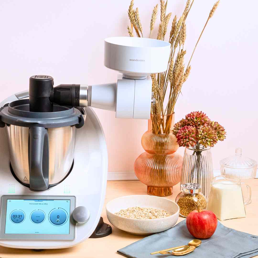 Grain Flaker mounted on a Thermomix in a styled kitchen with a bowl of oats, an apple and dried wheat