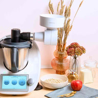 Grain Flaker mounted on a Thermomix in a styled kitchen with a bowl of oats, an apple and dried wheat