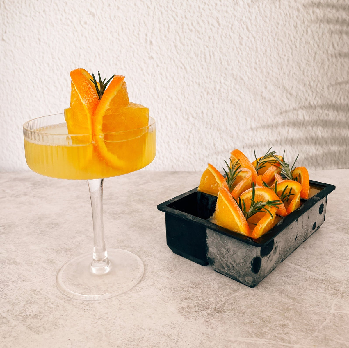Cocktail glass with a jumbo ice cube and orange garnish, beside a tray of frozen orange slices