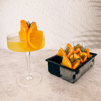 Cocktail glass with a jumbo ice cube and orange garnish, beside a tray of frozen orange slices