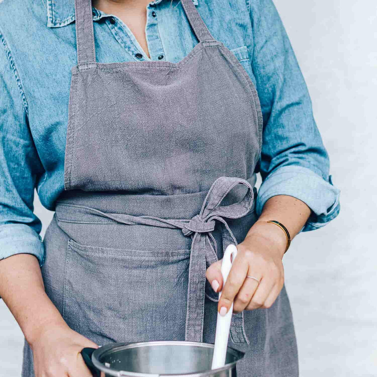 Close-up of the charcoal Linen Apron worn over a denim shirt, showing the cross-body straps and pocket