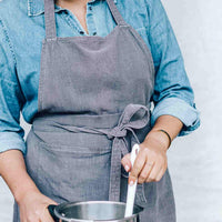 Close-up of the charcoal Linen Apron worn over a denim shirt, showing the cross-body straps and pocket