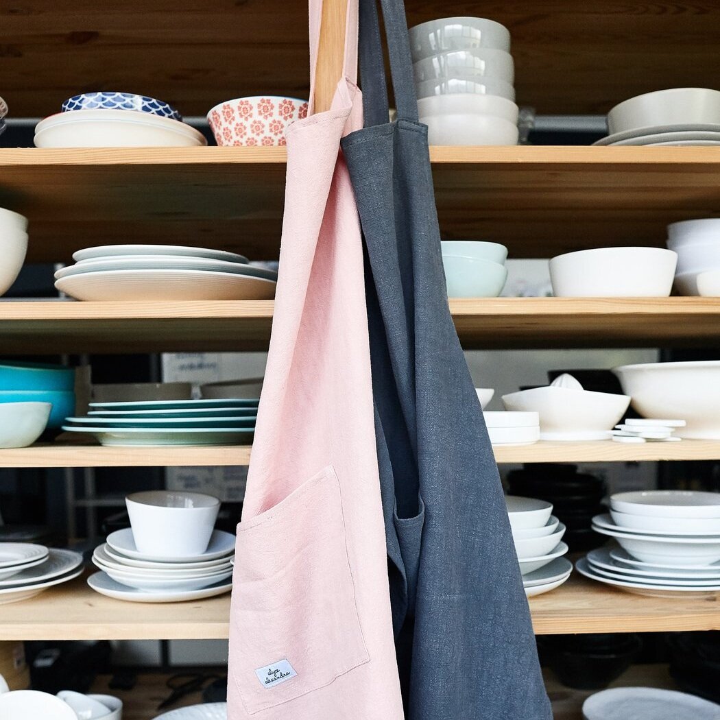 Charcoal and blush pink Linen Aprons hanging in front of open shelves of crockery