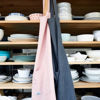 Charcoal and blush pink Linen Aprons hanging in front of open shelves of crockery
