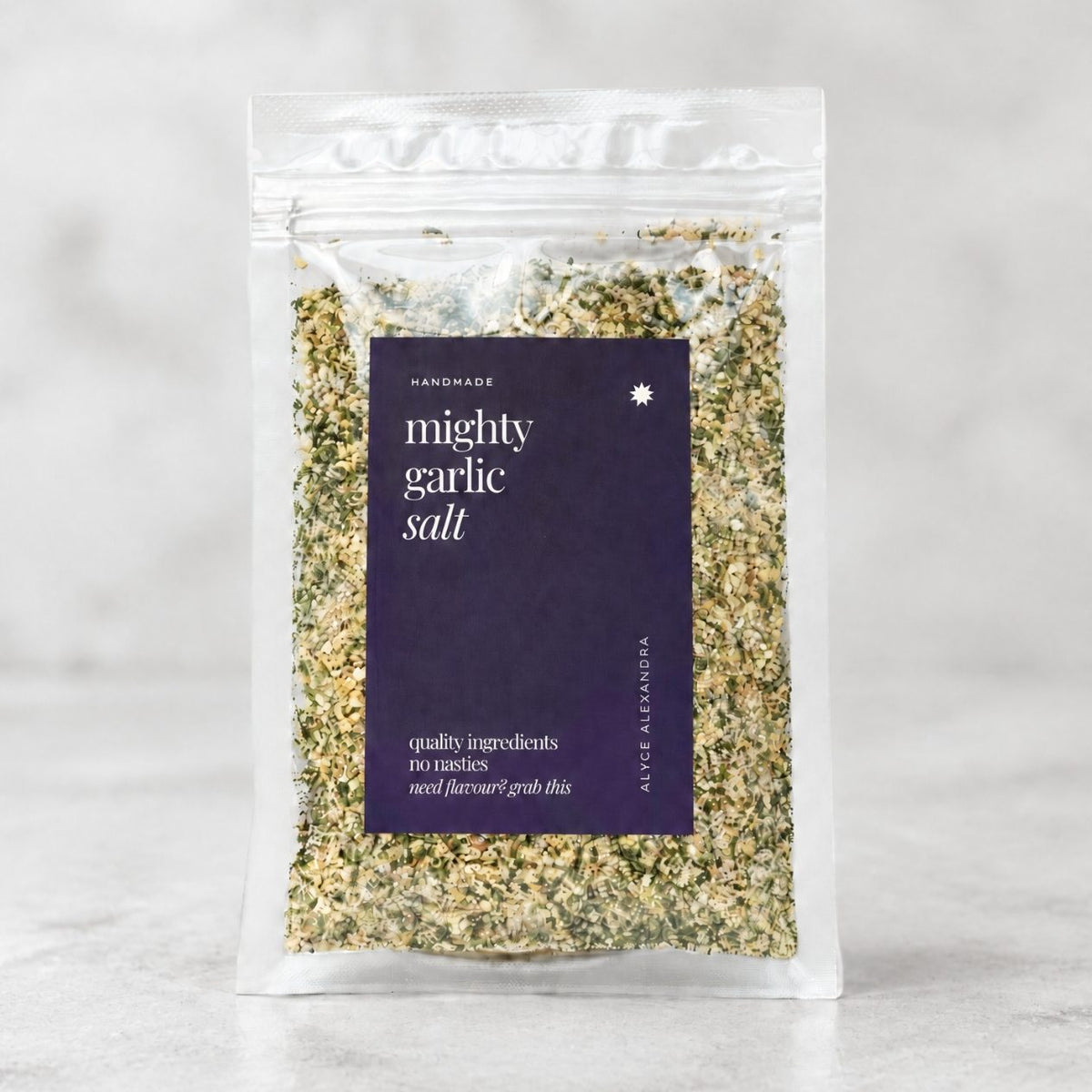 Alyce's Mighty Garlic Salt in a clear 100g packet with a deep purple label, the creamy garlic salt flecked with green parsley visible through the front