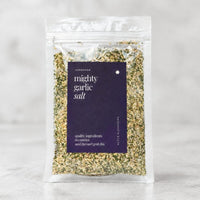Alyce's Mighty Garlic Salt in a clear 100g packet with a deep purple label, the creamy garlic salt flecked with green parsley visible through the front