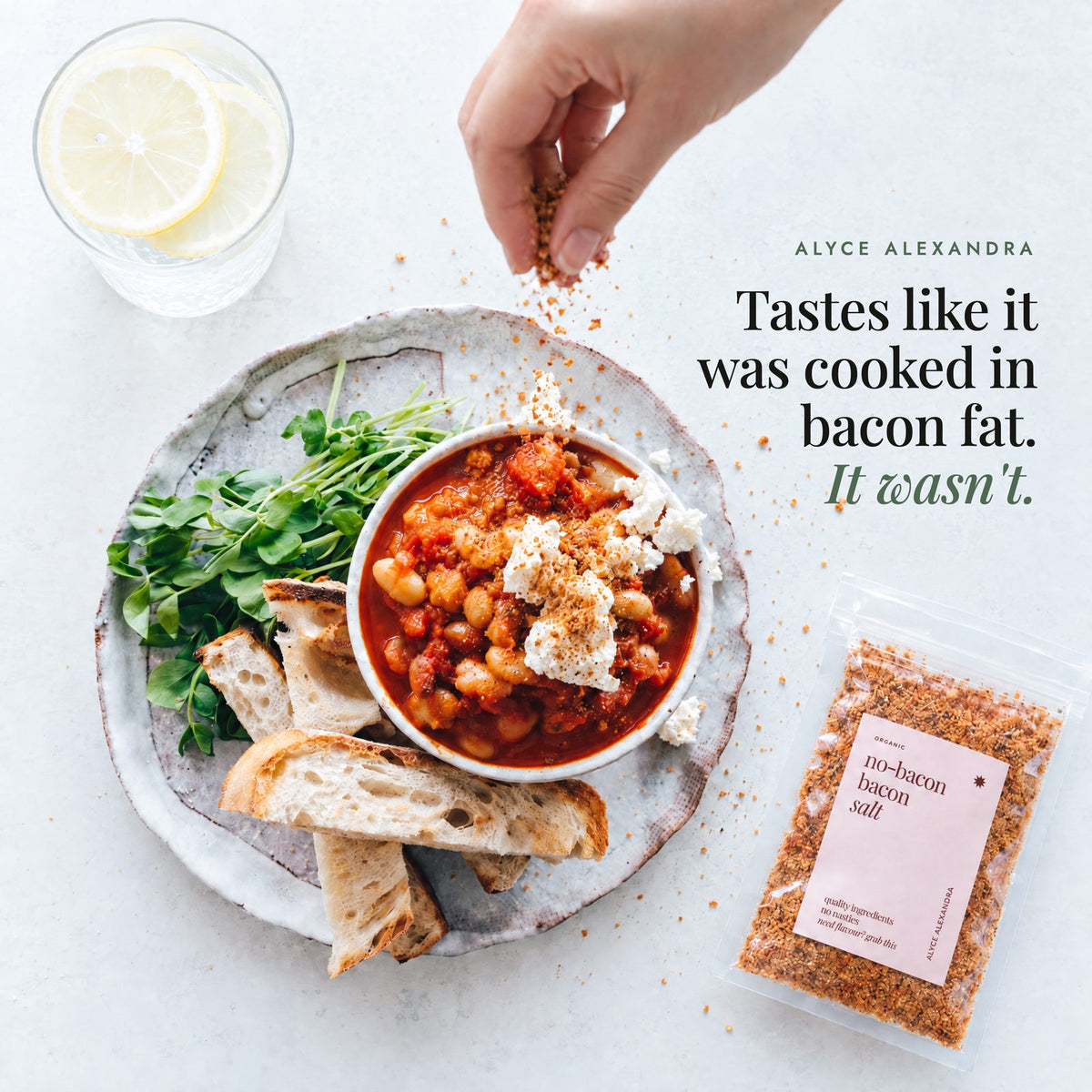 Hand sprinkling No-Bacon Bacon Salt over a bowl of tomato and white bean stew topped with crumbled feta, beside toasted bread, pea shoots, a glass of lemon water and a packet of the salt; tastes like it was cooked in bacon fat, it wasn't