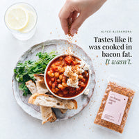 Hand sprinkling No-Bacon Bacon Salt over a bowl of tomato and white bean stew topped with crumbled feta, beside toasted bread, pea shoots, a glass of lemon water and a packet of the salt; tastes like it was cooked in bacon fat, it wasn't