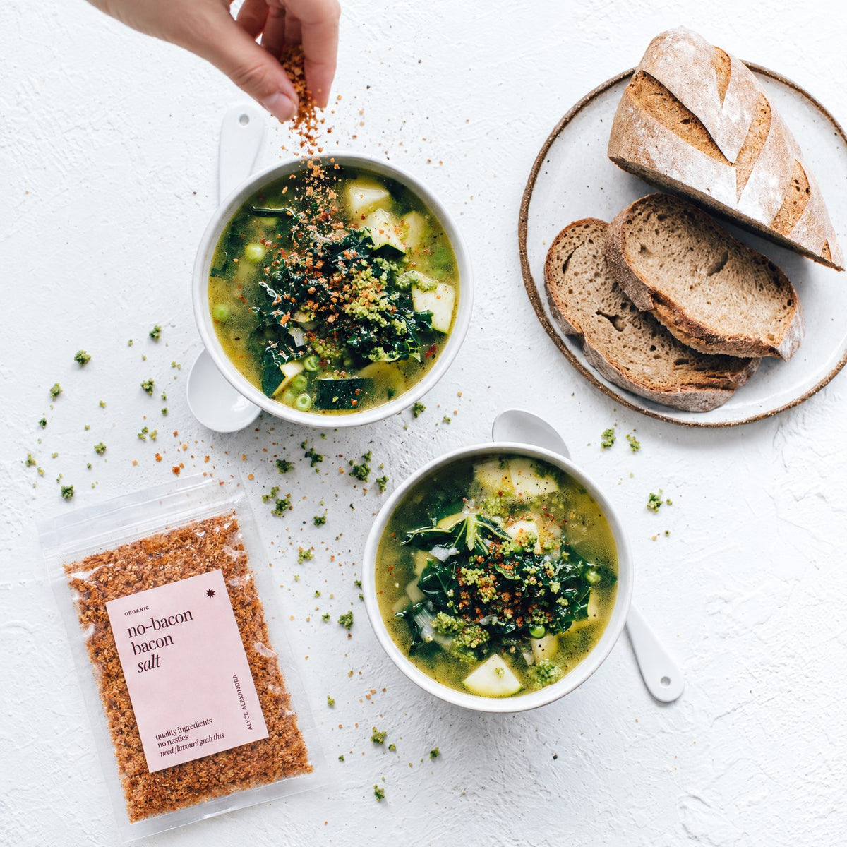 Hand sprinkling Organic No-Bacon Bacon Salt over two bowls of green vegetable soup with kale, zucchini and peas, beside sliced sourdough bread and a packet of the salt