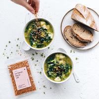 Hand sprinkling Organic No-Bacon Bacon Salt over two bowls of green vegetable soup with kale, zucchini and peas, beside sliced sourdough bread and a packet of the salt