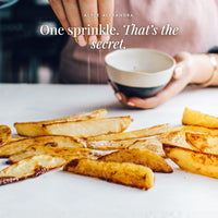 Hand sprinkling No-Bacon Bacon Salt from a small bowl over golden potato wedges; one sprinkle, that's the secret
