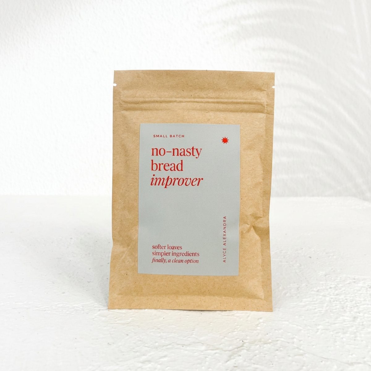 Kraft paper pouch of No-Nasty Bread Improver with its pale sage label reading softer loaves, simpler ingredients, standing on a white bench against a white wall